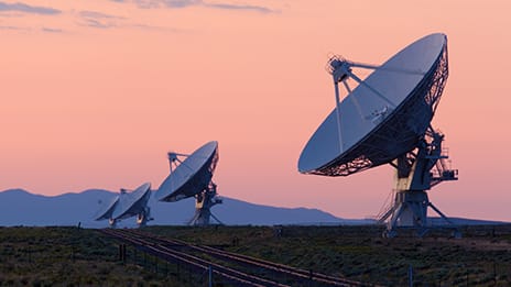 A row of satellite dishes against a pink sky with mountains in the background