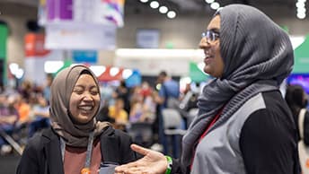 Two women in headscarves at a trade show; one of them gestures with her hands and the other one laughs