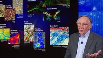 Jack Dangermond gestures with both hands while speaking next to a digital display of colorful maps