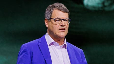 A presenter wearing glasses and a blazer against a dark green backdrop