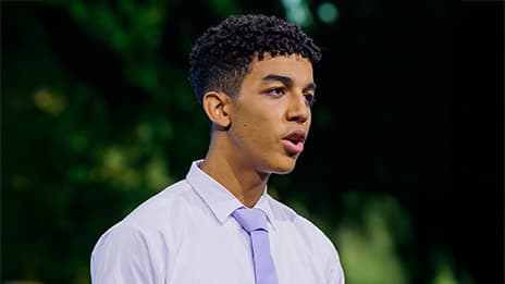 Youthful presenter wearing a lavender tie and dress shirt against a dark backdrop