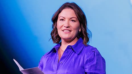 Brightly lit presenter with chin-length dark hair wearing a purple blouse against a blue backdrop