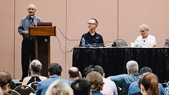 A presenter and a panel in a demo theater during a presentation, seen from behind the audience.