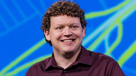 Smiling presenter with dark curly hair against a blue background with yellow lines