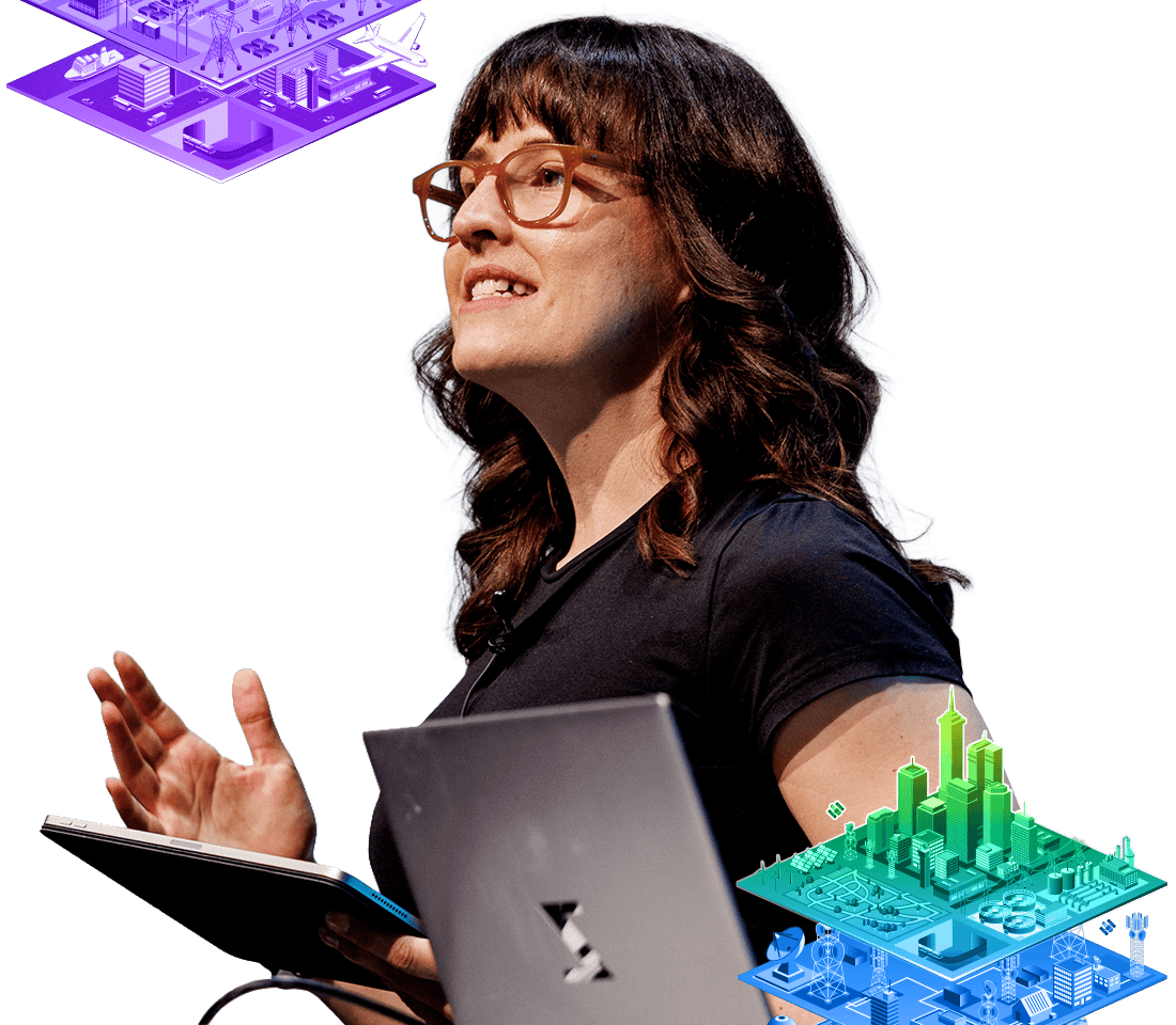 Presenter with long dark hair and glasses gestures while speaking against a map background