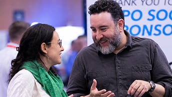 Bearded exhibitor gestures while bending towards an attendee wearing a green scarf
