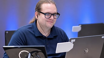 Person with glasses and a ponytail working at a laptop with another laptop to the left and more in the backgrground