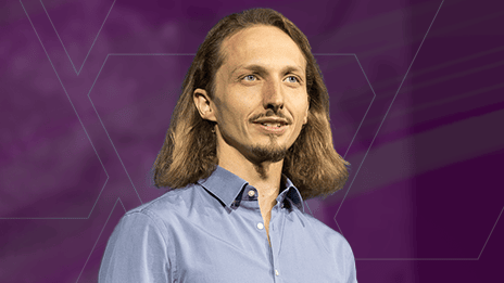 Closeup of a presenter with a manicured goatee and mustache and shoulder length hair on stage against a purple graphic background