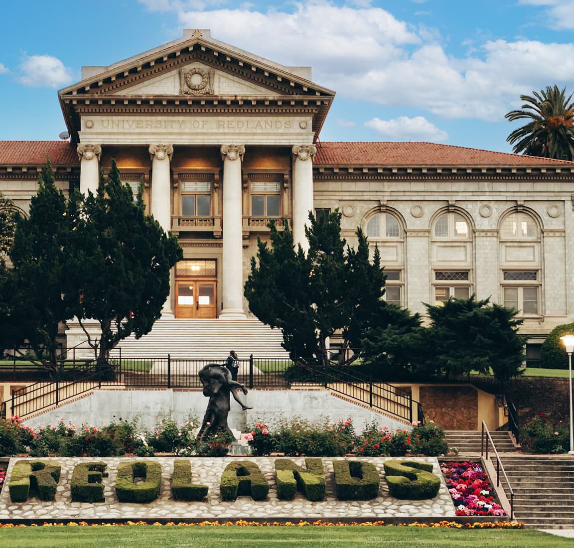 Historic University of Redlands building with grand columns and ornate architecture, framed by tall trees. In the foreground, a statue stands in front of manicured greenery spelling ‘REDLANDS.’