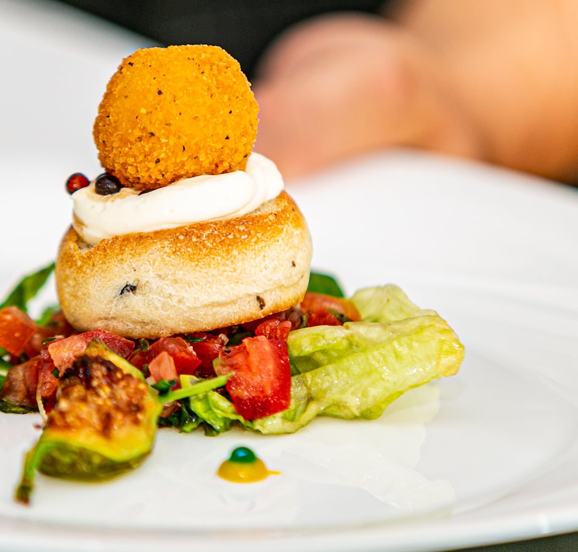 Close-up of a plated appetizer featuring a golden fried croquette on top of a toasted bread roll with a swirl of white cream, served over a fresh lettuce and diced tomato salad with a roasted Brussels sprout garnish