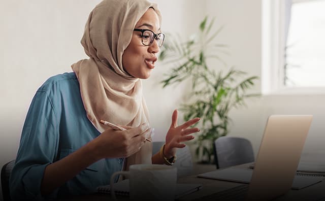 Woman wearing a hijab and glasses talks to colleagues on her laptop during a virtual conference meeting