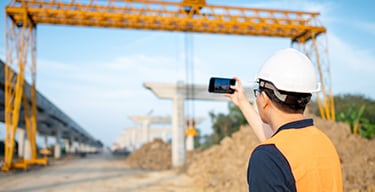Person in an orange vest and hard hat at a construction site, holding up a cell phone