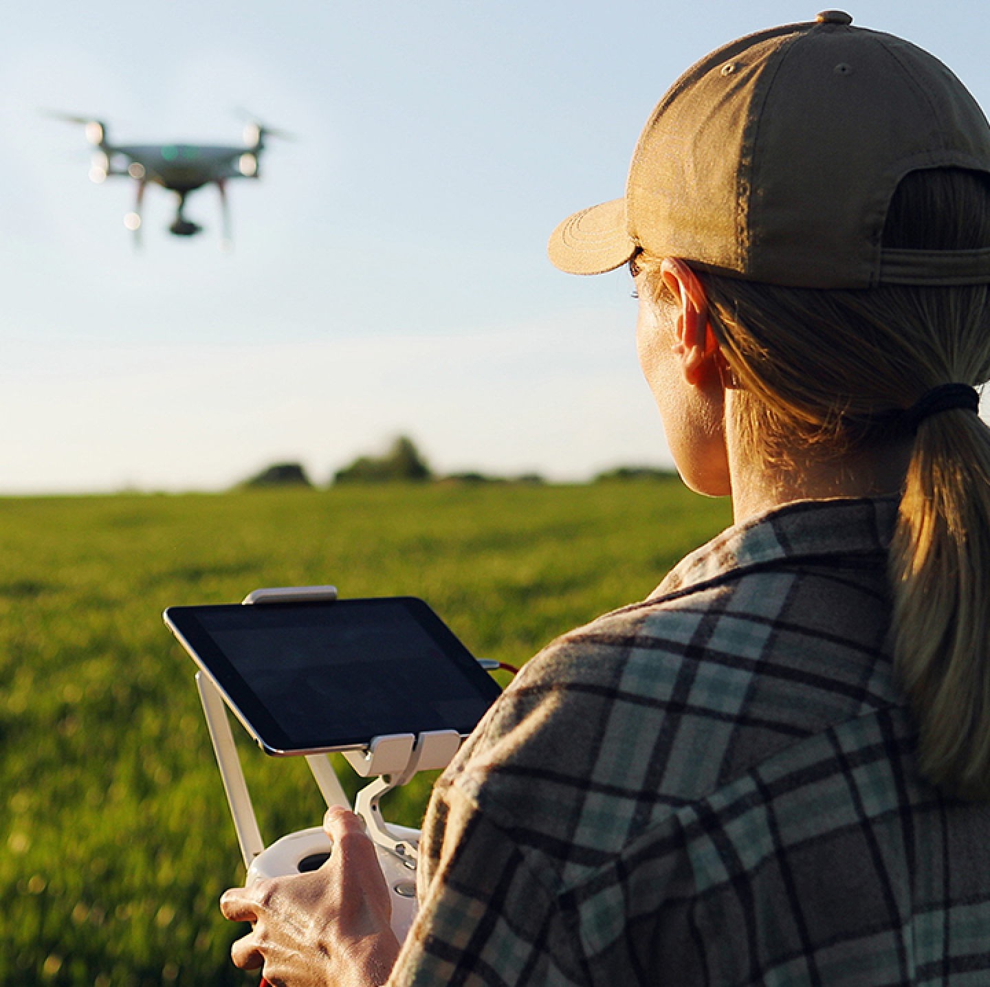 Over the shoulder view of a person in an open field wearing a plaid shirt and a hat while operating a drone
