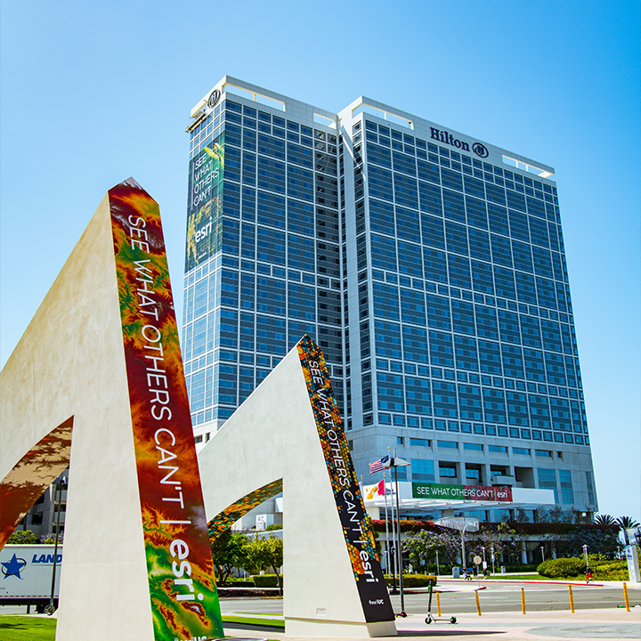 A photo of a gleaming blue glass hotel against a clear blue sky with colorful Esri event signage in front