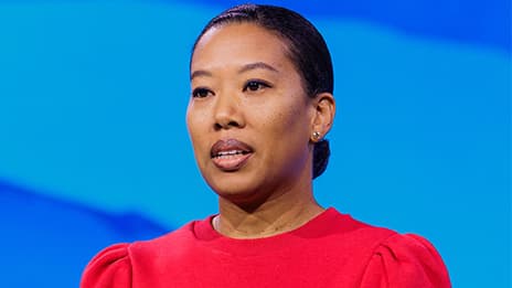 Closeup portrait of a presenter with their hair pulled back into a bun wearing a red cap-sleeved top against a light blue backdrop