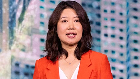 Closeup portrait of a presenter with their hair pulled back into a bun wearing a red cap-sleeved top against a light blue backdrop