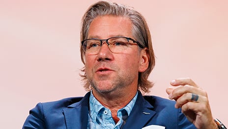 Speaker in a jacket and open-neck shirt gestures with their left hand while presenting
