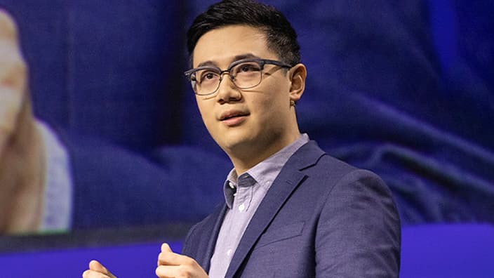 Close up of a conference speaker wearing glasses, a lavender dress shirt, and a dark blazer on stage during their presentation