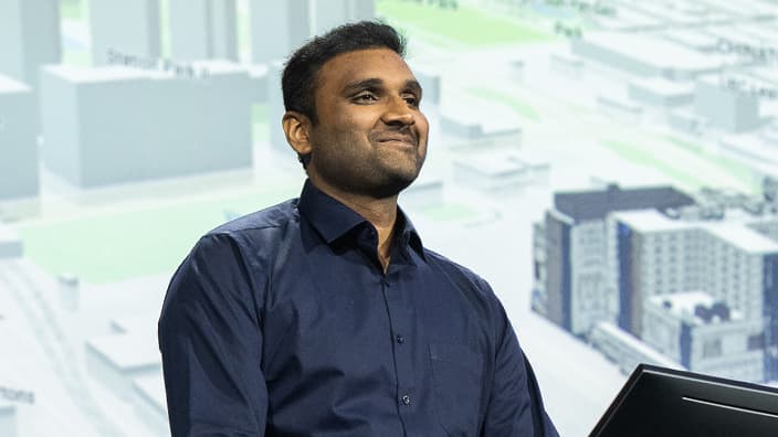 Smiling presenter, chin up, gazes towards an audience while standing against a blurry background showing a city rendering