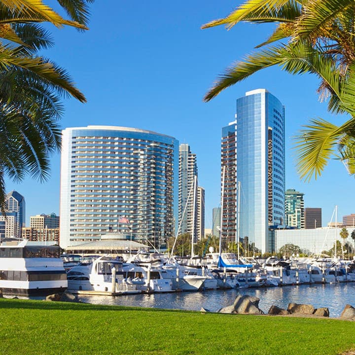 A photo of a gleaming silver glass hotel against a clear blue sky with a marina, grass, and palm trees in the foreground
