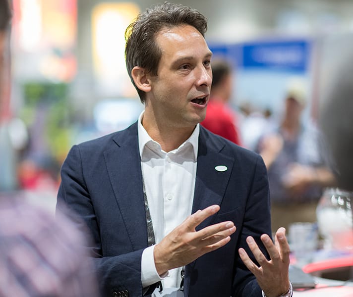 Person in a blue suit and open-neck white shirt gestures while talking to attendees at a trade show