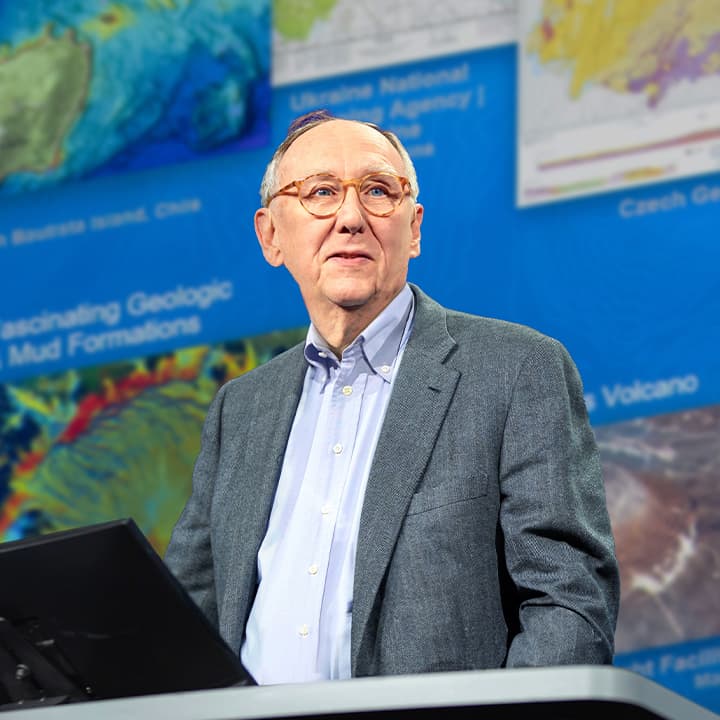 Jack Dangermond wearing a gray suit and round framed glasses during his Plenary Session presentation with colorful maps behind him.