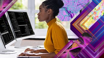 A focused developer wearing a mustard yellow sweater seated at a computer workstation with two monitors overlaid with colorful mapping graphics