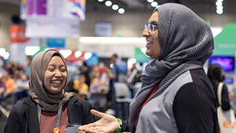 Two smiling attendees wearing headscarves laughing and interacting in the bustling Expo Hall