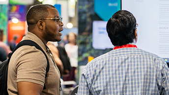 Side profile of an attendee engaged in conversation with an Esri expert wearing a red lanyard using a large monitor to display information