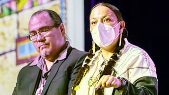 Two presenters standing side by side as one wearing a surgical mask and double braid hairstyle points in emphasis
