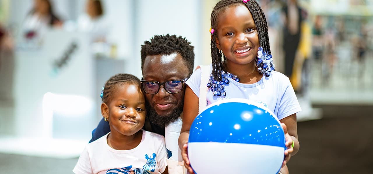 Father kneeling between his two smiling daughters as one holds a blue and white beach ball during a busy event