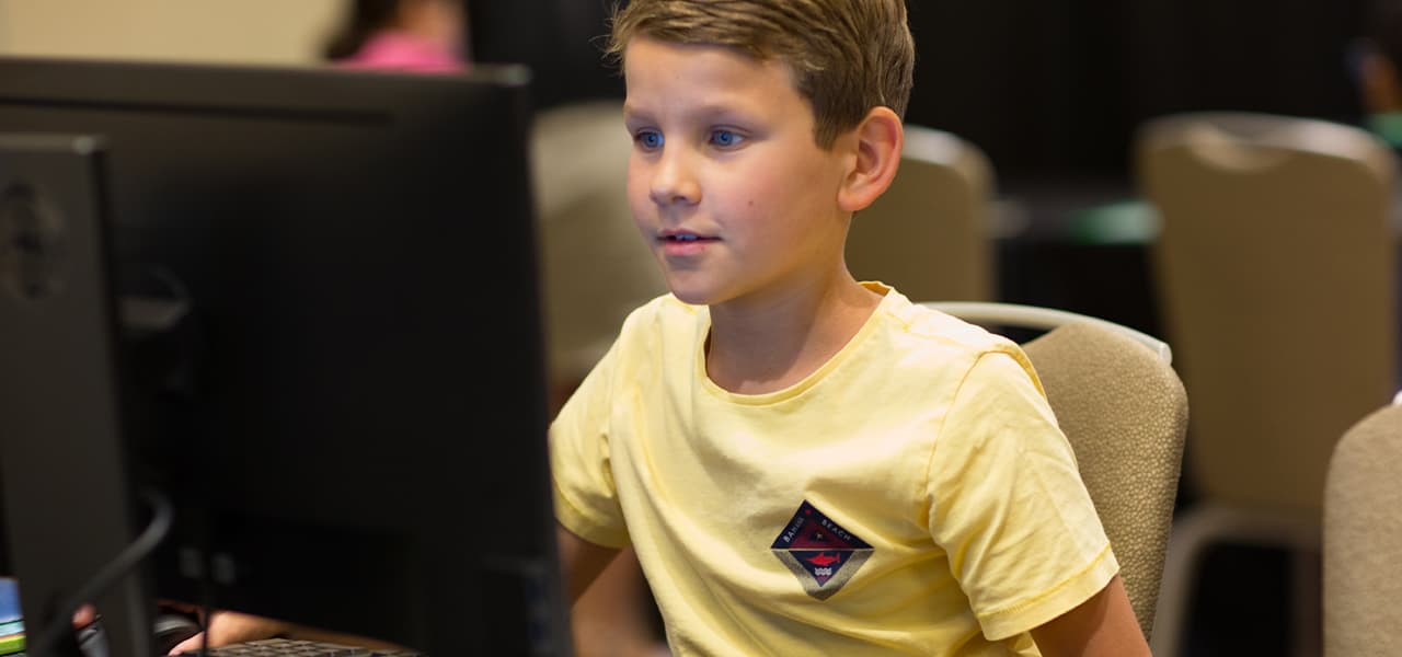 Child about ten years old wearing a yellow shirt sitting in a computer lab happily focused on a computer monitor