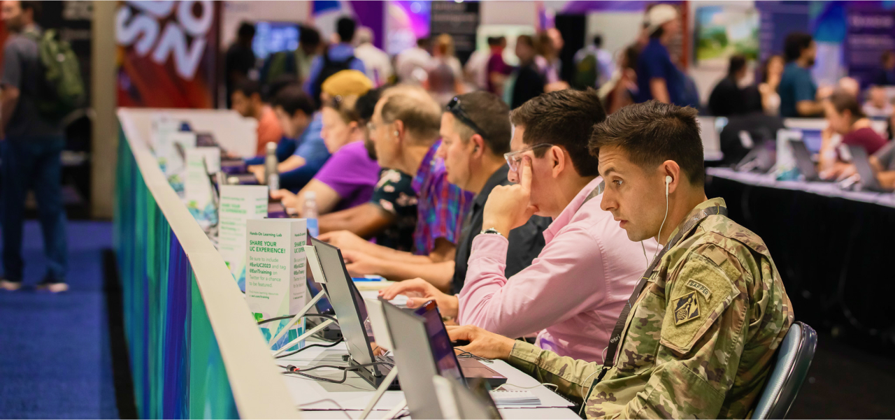 A group of Expo attendees sitting at a long table focused on laptops with one participant in a camouflage Army jacket seated at the near end