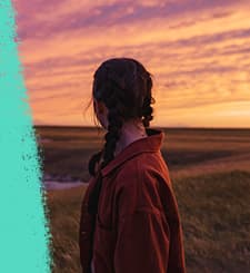 Person with braided hair wearing a jacket standing in an open field and looking toward a vibrant sunset with orange and purple clouds.