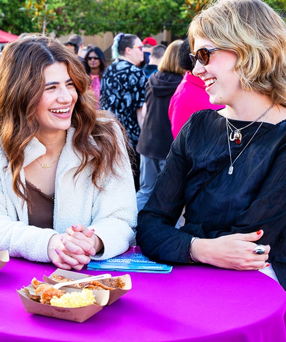 Two smiling people gathered outdoors at a table talk and enjoying food and drinks