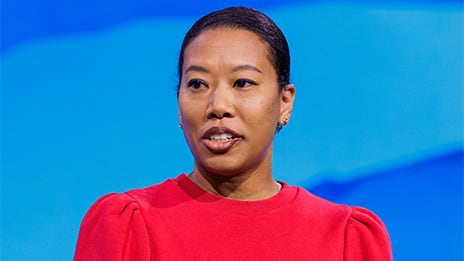 Person in a red top speaking on stage set against a smooth blue gradient background
