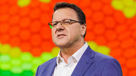 Person in glasses and a blue blazer speaking on stage in front of a bright orange and yellow hexagonal pattern