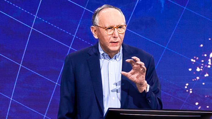 Jack Dangermond wearing a blue suit speaking on stage in front of a blue backdrop