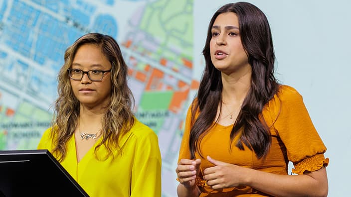 Two Esri experts presenting on stage in front of a large screen displaying a city map