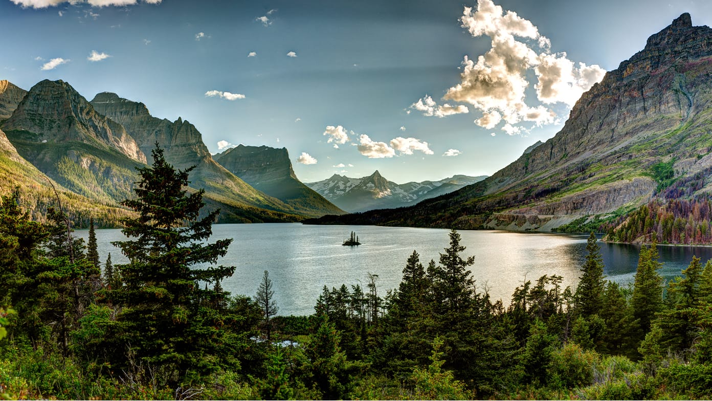 Panoramic view of a rippling lake surrounded by soaring rocky mountains with dense forest in the foreground
