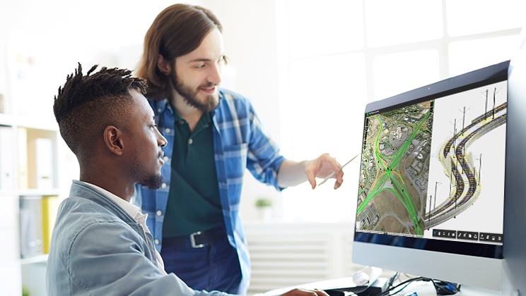 Zwei Personen in einem Büro, die auf einen Desktop-Computer schauen, auf dem ein geteilter Bildschirm zu sehen ist. Die eine Seite zeigt eine Baustelle mit grünen Linien und die andere Seite räumliche Daten zu einer Straße.
