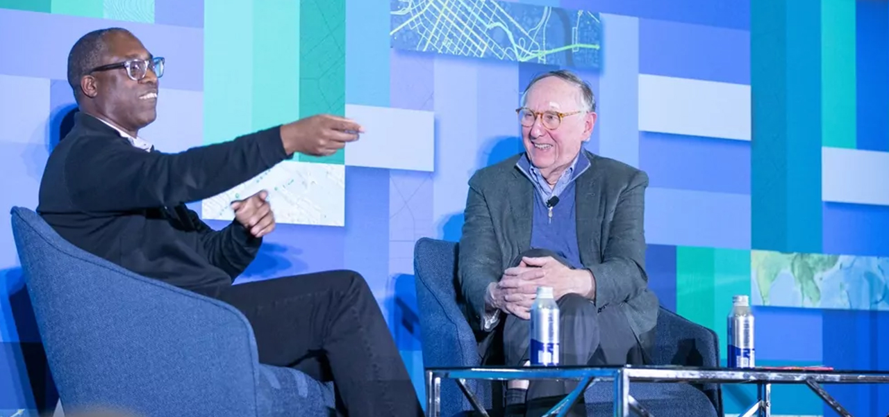 Photo of Jack Dangermond smiling and sitting with Kareem Yusef, Ph.D. onstage at a conference with an abstract background of soft purples and greens