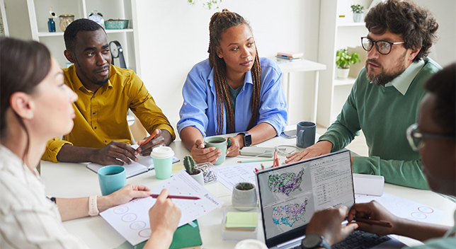 A group of business colleagues sitting around a table with their coffee cups, a laptop, pencils, and planning papers