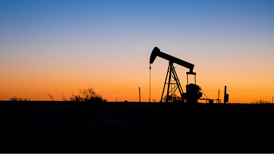 An oil rig in a broad flat field silhouetted against a pale orange sunset
