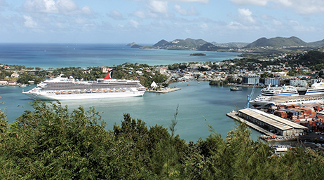 The coast of a lush seaside city with cruise ships and boats in a marina, vast waters beyond, and green hills in the distance