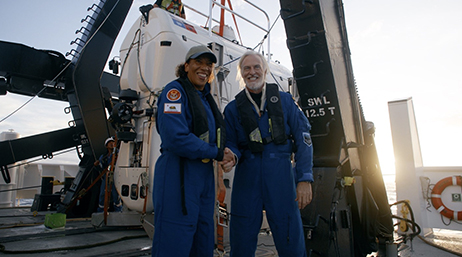 Esri Chief Scientist Dawn Wright shaking hands with Victor Vescovo on the deck of a ship while a diving vessel hangs from a crane