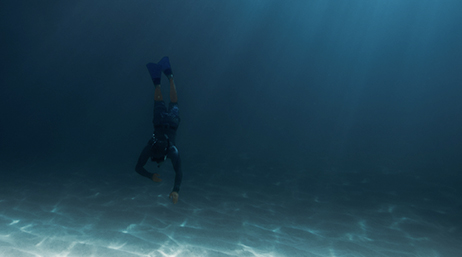A person wearing a snorkel, goggles, and swimming fins diving toward the ocean floor as sunlight filters through the water