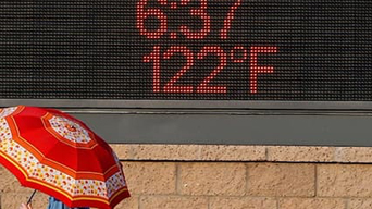 A person shaded by a red umbrella walking in front of a sign that has the time and temperature displayed