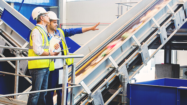 Two people in yellow safety vests and hard hats discuss a slanting conveyor belt in a clean white warehouse