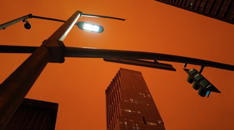 A view of a tall city building, a streetlight, and a traffic light with a dark orange sky in the background